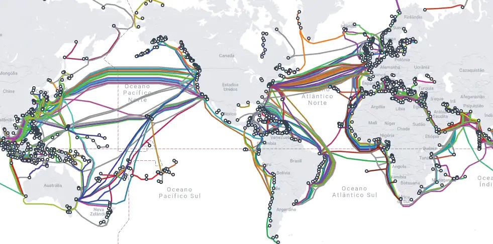 Planisfério centrado no Oceano Pacífico com centenas de linhas coloridas representando os cabos submarinos de fibra óptica que cruzam os oceanos e conectam os continentes. Os traçados se concentram no Atlântico Norte, entre a América do Norte e a Europa; no Pacífico Norte, entre a Ásia e a América do Norte; e ao longo das costas da América do Sul, da África e do sul da Ásia. Pequenos círculos marcam os pontos onde os cabos chegam ao litoral, como no Nordeste brasileiro, no leste da Ásia e no entorno da Europa.